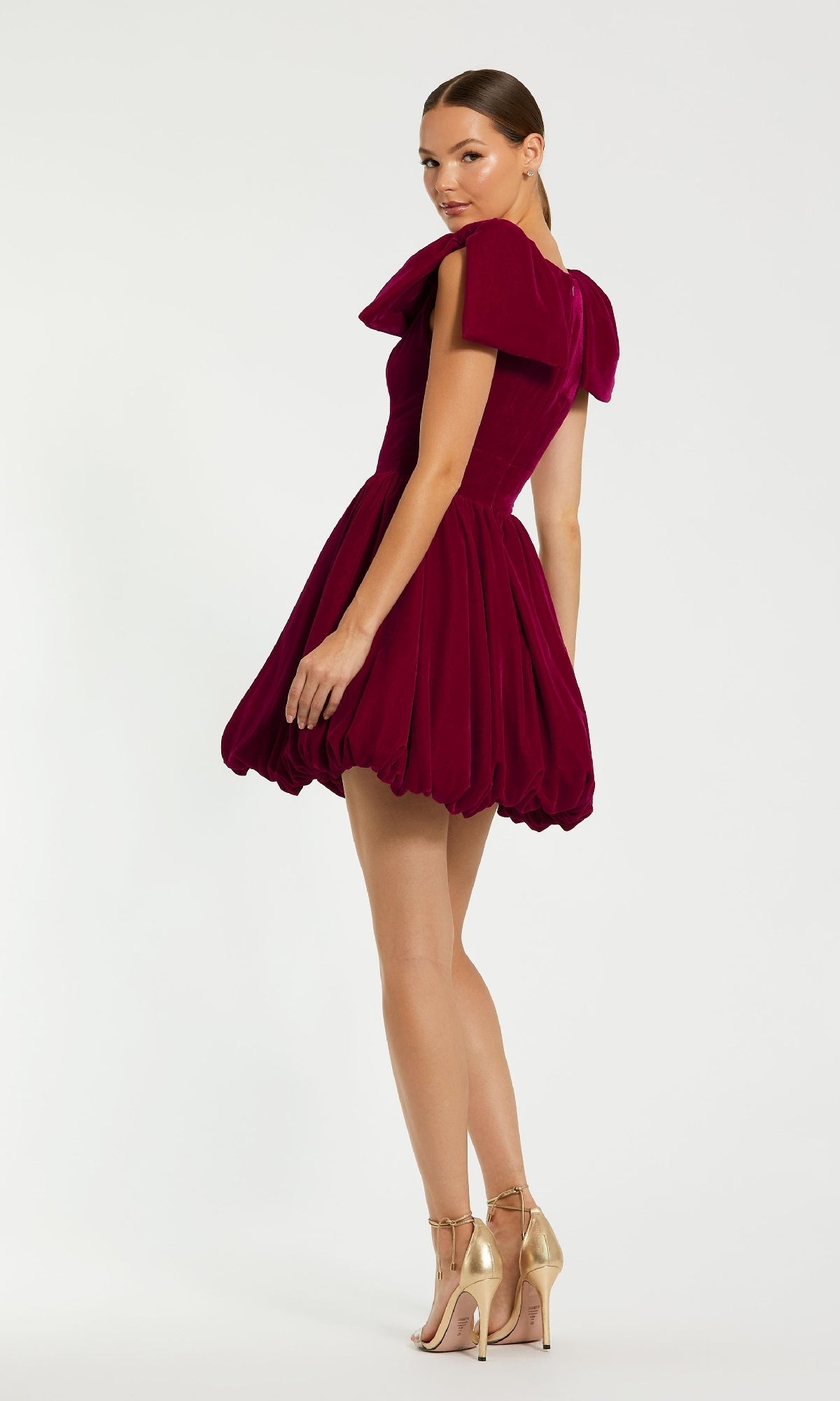 A woman wears the Mac Duggal 11743 Berry Red Mini Semi-Formal Dress with puffed sleeves and a bubble hem, paired with gold high-heeled sandals, standing against a plain white background.